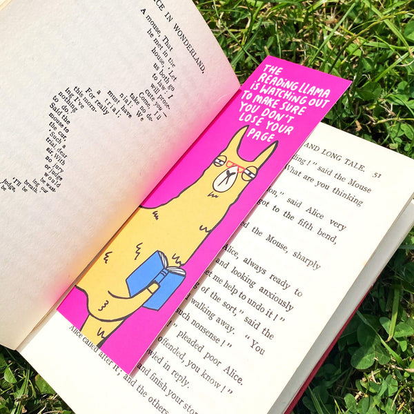 A bookmark with a llama illustration by Katie Abey. They have glasses on and are reading a book. Text above says The reading llama is watching out to make sure you don't lose your page. It has a pink background It is inside a book