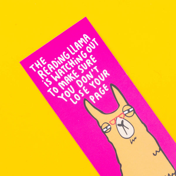 A bookmark with a llama illustration by Katie Abey. They have glasses on and are reading a book. Text above says The reading llama is watching out to make sure you don't lose your page. It has a pink background