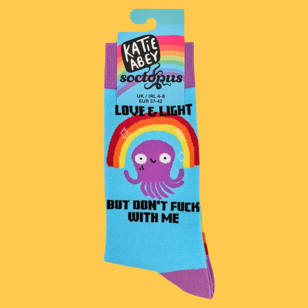 A close-up of Katie Abey's "Love & Light But Don't F**k With Me" socks, featuring a purple octopus under a rainbow. The packaging highlights the brand's whimsical charm, celebrating individuality with these colourful and bold socks designed to brighten your day.