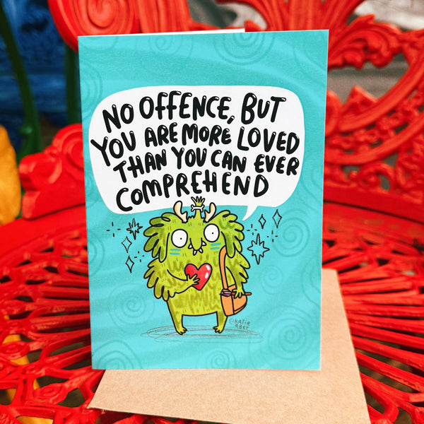 A6 illustrated greeting card by Katie Abey featuring a cute green fluffy monster holding a red heart and wearing a tiny crown, with a speech bubble that reads “No offence, but you are more loved than you can ever comprehend”. The card has a turquoise background with swirl details and is photographed standing upright with a kraft envelope behind it.