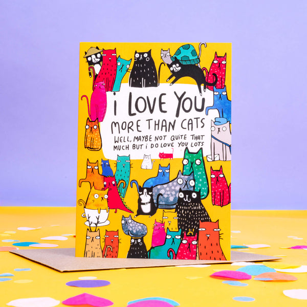 I love you more than cats a6 greeting card stood up on the brown envelope the card comes with, silver glittery paper and love hearts sweets on a white background. The card cover is a yellow background with various different coloured, patterned and sized cats with text in the top centre reading 'I love you more than cats. Well, maybe not quite that much but I do love you lots' Designed by Katie Abey in the UK.