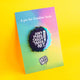 Katie Abey “Don’t Make Sense Make Art” pin badge displayed on a pastel rainbow backing card with the phrase “A pin for creative souls”. The enamel pin features white text and a pencil illustration, finished in iridescent metal on a bright yellow background.