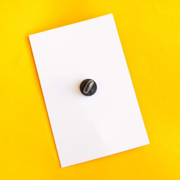 Back view of the Katie Abey enamel pin badge showing a black rubber clutch fastening on a white display card, placed on a vibrant yellow background. The pin is secured and designed for easy attachment to jackets, bags or accessories.