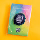 Katie Abey enamel pin badge packaged in a clear sleeve on a colourful gradient backing card reading “A pin for creative souls”. The rainbow finish pin with white lettering and pencil design stands out against a bold yellow background.
