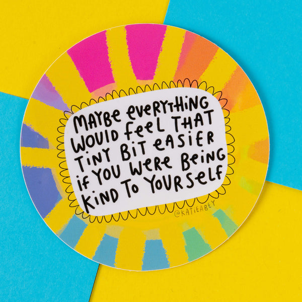 A circular sticker from Katie Abey with the message, "Maybe everything would feel that tiny bit easier if you were being kind to yourself." It showcases a bold, multi-coloured sunburst background in shades of yellow, pink, blue, and orange. The quirky design is part of Katie Abey's mission to spread positivity and celebrate individuality.