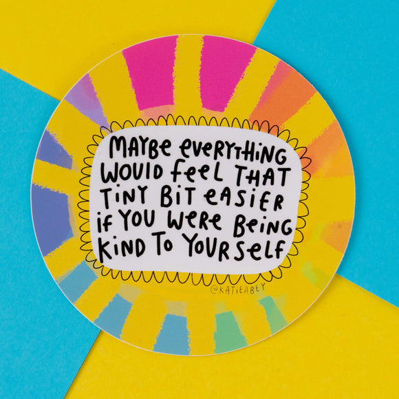 A round, colourful sticker by illustrator Katie Abey, featuring the text "Maybe everything would feel that tiny bit easier if you were being kind to yourself." The design has a vibrant, rainbow sunburst pattern with yellow, pink, blue, and orange accents. Katie Abey's unique style encourages self-kindness and embracing your weirdness.