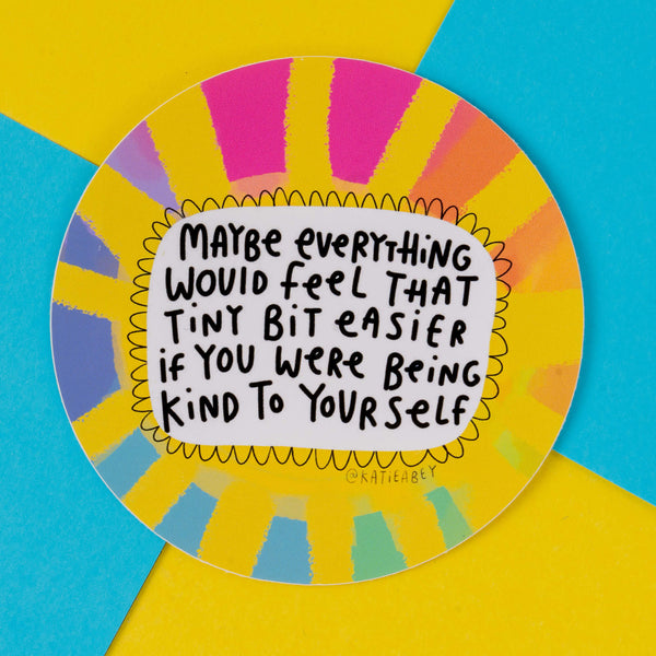 A round, colourful sticker by illustrator Katie Abey, featuring the text "Maybe everything would feel that tiny bit easier if you were being kind to yourself." The design has a vibrant, rainbow sunburst pattern with yellow, pink, blue, and orange accents. Katie Abey's unique style encourages self-kindness and embracing your weirdness.