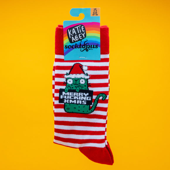 Katie Abey Sweary Cat Merry F*cking Christmas Socks displayed in packaging. The red and white striped socks feature a green cat in a Santa hat holding the sweary festive message. Bright yellow background. Fun novelty Christmas socks by UK illustrator Katie Abey.