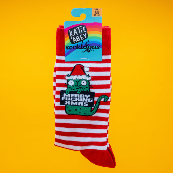 Katie Abey Sweary Cat Merry F*cking Christmas Socks displayed in packaging. The red and white striped socks feature a green cat in a Santa hat holding the sweary festive message. Bright yellow background. Fun novelty Christmas socks by UK illustrator Katie Abey.