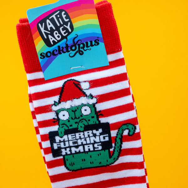 Close up of Katie Abey’s Sweary Cat Merry F*cking Christmas Socks, showing the green cat in a Santa hat on red and white stripes. The colourful Katie Abey Socktopus label is attached. Festive novelty socks by UK illustrator Katie Abey, photographed on a bright yellow background.