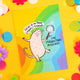 Fun cute A6 silk finish greeting postcard, featuring illustrated happy molerat holding magnifying glass, on colourful rainbow background. Reading 'The magnifying molerat' and 'focus in on those sparkly moments' in black letters and speech bubble. Designed by Katie Abey in the UK.