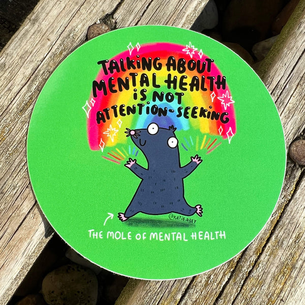 Green circular sticker of a mole with rainbows coming from its fingertips with text above saying talking about mental health is not attention seeking