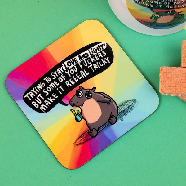 Katie Abey Moo Deng square coaster with a bold design of a sassy hippo holding a lighter, and the message, "Trying to stay 'love and light' but some of you fuckers make it reeaal tricky." Set against a vibrant rainbow gradient, this piece brings colour and attitude to your table. Katie Abey’s unique illustrations are made to inspire embracing the weird side in everyone. Ideal for quirky home decor lovers and fans of alternative style.