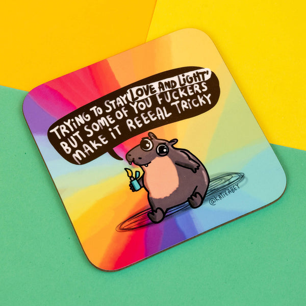 Katie Abey Moo Deng square coaster with a humorous, slightly edgy design. Features a hippo holding a lighter, saying, "Trying to stay 'love and light' but some of you fuckers make it reeaal tricky." Set against a rainbow gradient background, this item adds personality to any space. Perfect for alternative decor fans and those who love a bit of dark humour. Katie Abey’s work inspires embracing the quirky side of life.