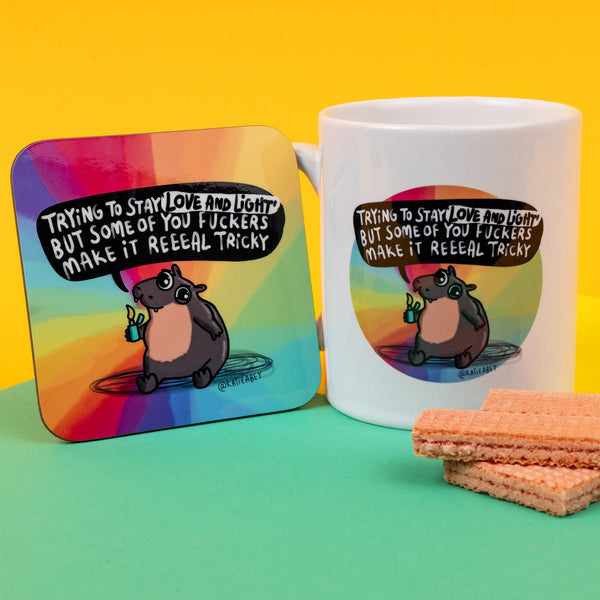 Katie Abey Moo Deng coaster and mug combo, showcasing a cheeky hippo holding a lighter, saying, "Trying to stay 'love and light' but some of you fuckers make it reeaal tricky." The rainbow gradient background adds charm to this quirky illustration. Perfect for alternative home decor or gift ideas that celebrate individuality. Katie Abey's designs encourage a playful and weird approach to life, perfect for those who enjoy a bit of sass.