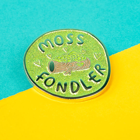The Moss Fondler Enamel Pin Badge on a blue and yellow background. The glittery green circle enamel pin has a shiny gold coloured outline with a brown log in the centre covered in moss with hearts and text reading 'moss fondler' surrounding. Hand drawn design by Katie Abey for nature lovers.