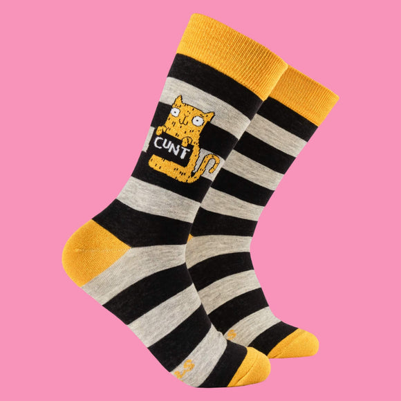 Bold, striped socks featuring black and grey bands, yellow accents, and a quirky orange cat holding a cheeky sign reading "C**T." Designed by Katie Abey, these socks bring humour and weirdness to your wardrobe. Perfect for quirky gift-givers or fun sock enthusiasts.