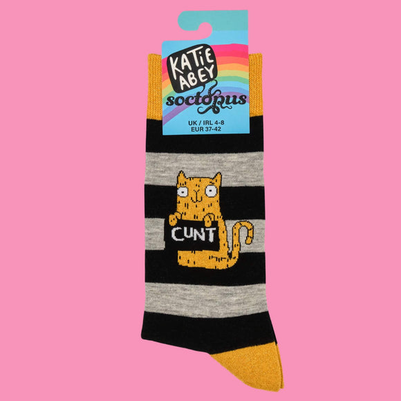 A close-up of Katie Abey’s “C**T Cat” socks, with playful black and grey stripes, yellow detailing, and a sassy orange cat illustration. Packaged in vibrant Katie Abey branding. Made to inject personality and fun into everyday style for weirdness-lovers.