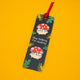 Angled view of Katie Abey mushroom bookmark with red ribbon tassel, showing “Stop talking, I’m reading” text and cute toadstool illustration. Dark green leafy design on bold yellow background. Perfect fun gift for bookworms and lovers of quirky stationery.