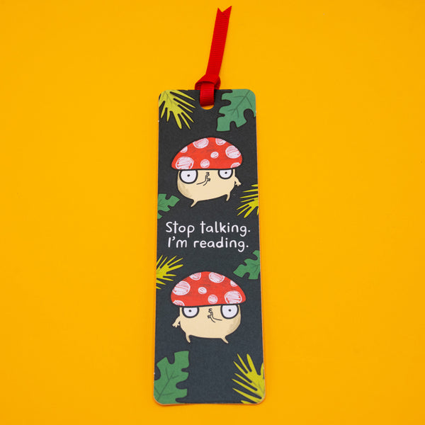 Full view of Katie Abey “Stop Talking, I’m Reading” mushroom bookmark with red ribbon tassel on bright yellow background. Features playful toadstool characters and leafy pattern. Ideal quirky gift for readers, book lovers and fans of fun illustrated stationery.
