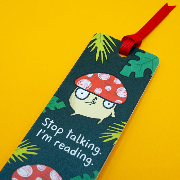 Close-up of Katie Abey mushroom bookmark featuring quirky red toadstool character and text “Stop talking, I’m reading” on dark green leafy background. Red ribbon tassel visible. Fun illustrated bookmark for readers, bookworms and lovers of cute, weird designs.

