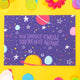 blue postcard with planets and stars drawn on with the words - you should know you are not alone written in the middle. It is on a yellow background with confetti, pom poms and fake daisies in different colours