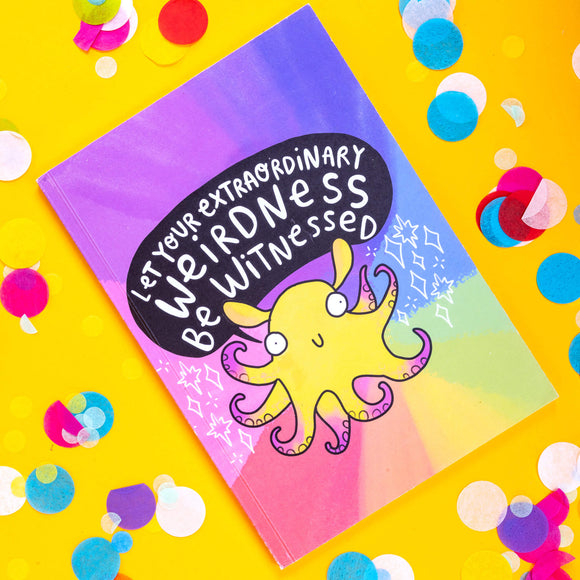 a rainbow coloured notebook with a yellow octopus illustration with purple tentacles and a smiling face on it. The octopus has a speech bubble above it's head that reads 'let your extraordinary weirdness be witnessed' and white stars around it. The notebook is lying on a yellow surface.