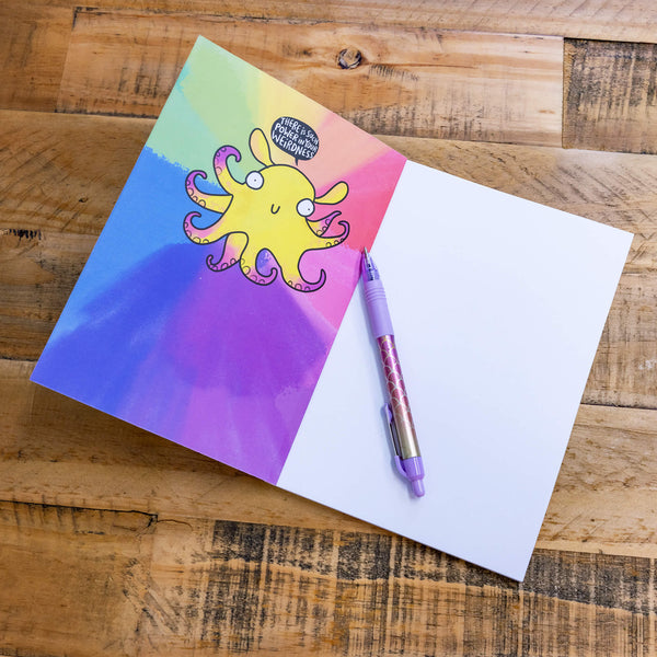 a rainbow coloured notebook with a yellow octopus illustration with purple tentacles and a smiling face on it. The octopus has a speech bubble above it's head that reads 'let your extraordinary weirdness be witnessed' and white stars around it. The notebook is lying on a yellow surface.