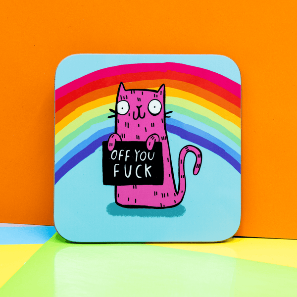 A blue coaster and rainbow coaster with a pink illustrated cat by Katie Abey holding a sign saying 'Off you fuck' it is leaning against a colourful background