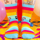a model with tattooed legs wearing Katie Abey off you fuck socks with pink cat holding a sign. They are rainbow striped and lovely and vibrant. The model is stood on a yellow floor with balloons, confetti and ribbons
