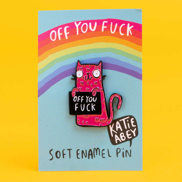 Katie Abey enamel pin on a display card, featuring a pink cat with bulging eyes and a cheeky grin. The cat holds a sign with the phrase 'Off You Fuck', set against a pastel blue background with a rainbow arching overhead