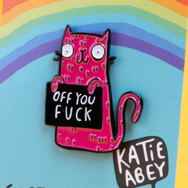 Katie Abey pink cat enamel pin featuring a quirky, wide-eyed cat holding a black sign with the bold message 'Off You Fuck'. The pin is displayed against a vibrant, rainbow background on a branded card