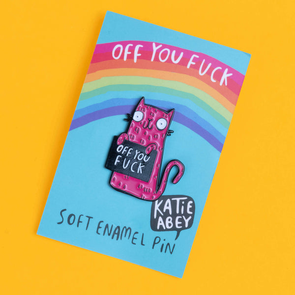 Soft enamel pin by Katie Abey, featuring a pink cat character holding a sign with the humorous phrase 'Off You Fuck'. The pin is mounted on a branded card with a rainbow design and displayed against a solid yellow backdrop.