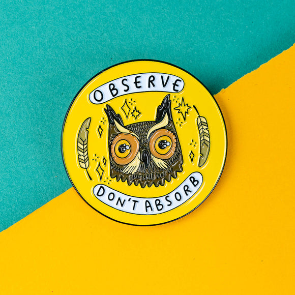 The Observe Don't Absorb Enamel Pin Badge on a yellow and teal card background. The yellow base circle soft enamel pin has a brown smiley owl with big eyes surrounded by sparkles and two feathers with text in white banners reading 'observe don't absorb'.