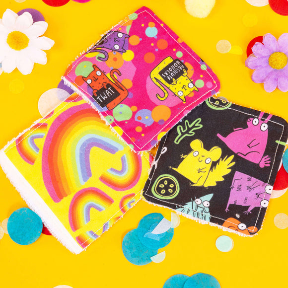 colourful square shaped reusable makeup remover pads with Katie Abey characters and illustrations on them. They are laying on top of a bright yellow backdrop with coloured confetti and flowers scattered around