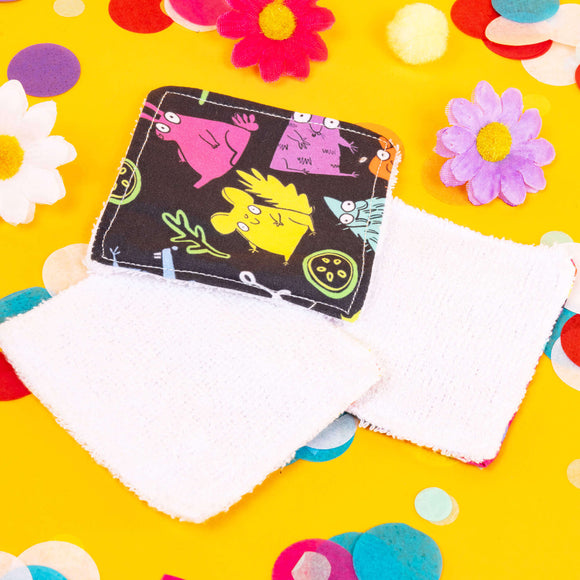 a dark grey square shaped makeup remover pad with colourful Katie Abey character illustrations on it. There are two pads next to it that are laid face down to reveal the plain white side of the makeup remover pads. They are laying on top of a bright yellow backdrop with coloured confetti and flowers scattered around