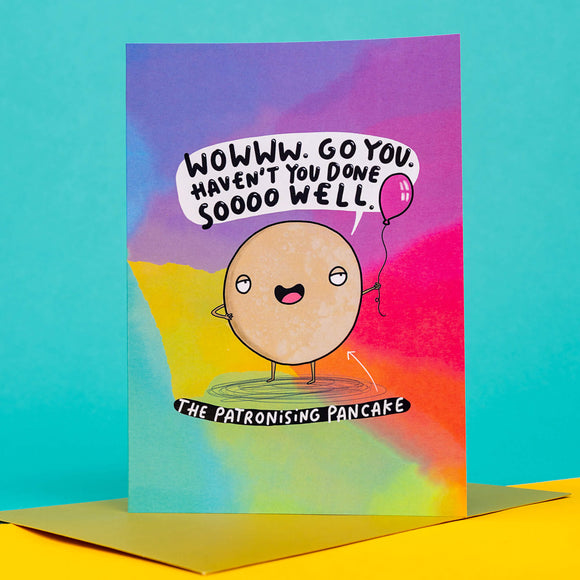 funny congratulations A6 card featuring The Patronising Pancake holding a purple balloon with a smug expression and speech bubble that reads 'wowww. Go you. Haven't you donesoooo well.' on a rainbow coloured card with pairing gold envelope. Designed by Katie Abey.