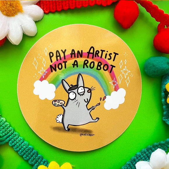 A sticker on a green background with a drawing by Katie Abey of a rabbit with a paint brush and a rainbow behind it with text saying pay an artist not a robot