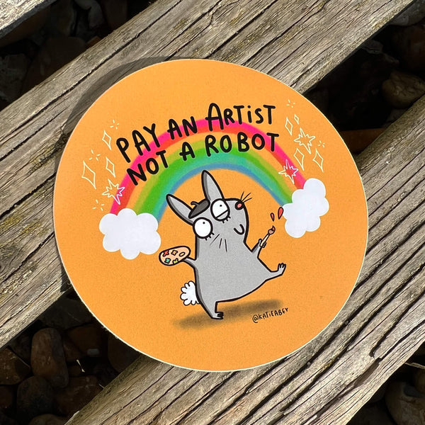 A sticker on wooden decking with a drawing by Katie Abey of a rabbit with a paint brush and a rainbow behind it with text saying pay an artist not a robot