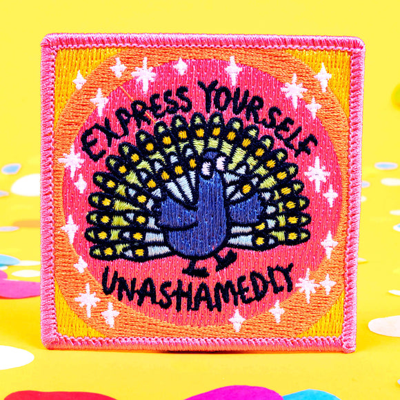 A patch with a peach background and a peacock illustration by Katie Abey in embroidery with Express yourself unashamedly written around it