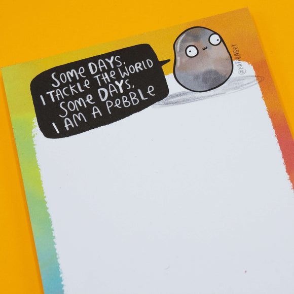 Close-up of the Katie Abey "Some Days I Am A Pebble Pad" notepad featuring a cute illustrated pebble saying "Some days I tackle the world, some days I am a pebble" on a rainbow border background. Fun stationery gift from small business Katie Abey.