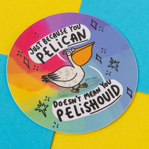 A round sticker designed by Katie Abey showing a humorous illustration of a pelican, set against a vibrant rainbow gradient background. The text humorously states, "Just because you PELICAN doesn’t mean you PELISHOULD." This design reflects the illustrator’s signature theme of celebrating individuality and embracing weirdness.