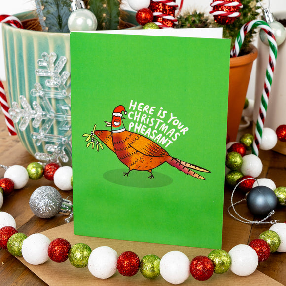 A bright green Christmas card featuring a cartoon pheasant wearing a Santa hat and holding mistletoe. The text says, "Here is your Christmas pheasant." The card is surrounded by festive baubles and holiday decorations.