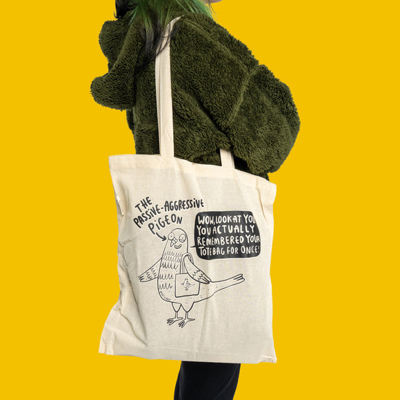 Model wearing Passive Aggressive Pigeon Tote Bag by Katie Abey over the shoulder, styled with a green textured jacket. Reusable cotton canvas tote with funny pigeon illustration and witty slogan, ideal eco shopping bag with quirky alternative style from an independent UK illustrator.