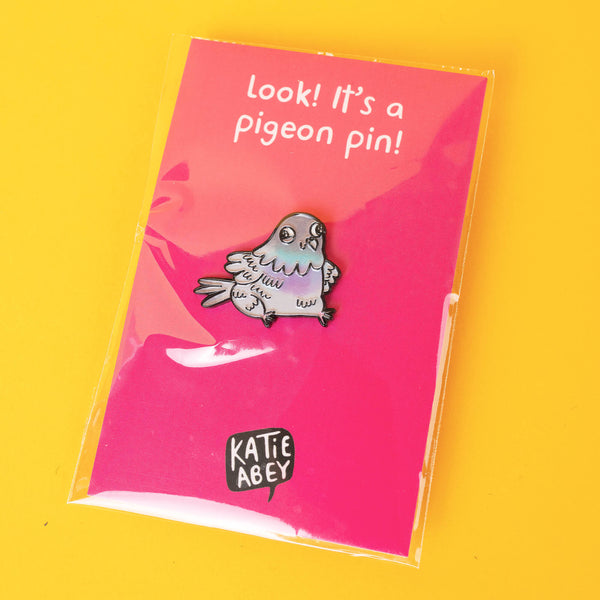 Katie Abey pigeon enamel pin in protective packaging on a bright pink backing card that says, "Look! It's a pigeon pin!" This quirky, pastel pigeon design is ideal for lovers of cute, eccentric accessories or collectors of unique pins.