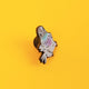 A close-up of Katie Abey's pigeon enamel pin resting on a yellow backdrop, showcasing its unique pastel hues and metallic shimmer. Designed to inspire joy and embrace your weird side, this pigeon pin adds a quirky flair to any outfit or collection.