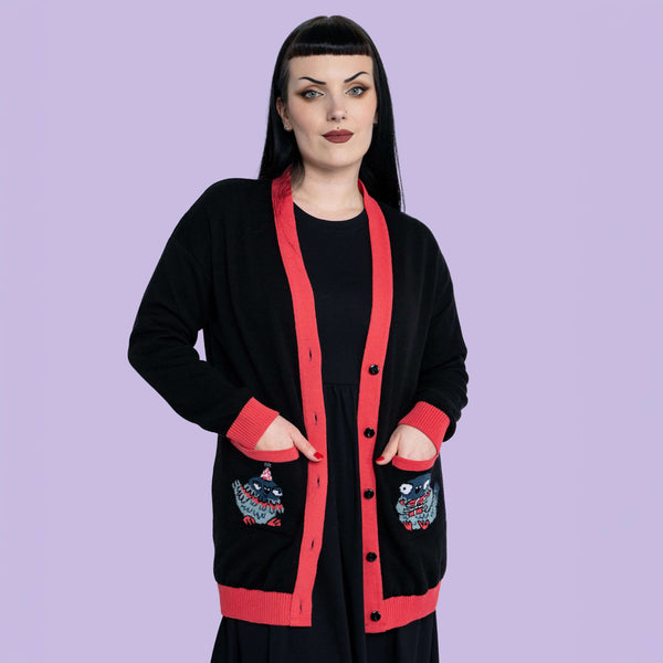 Model wearing Katie Abey x Rebel Romance Pigeon Party Long Cardigan styled over a black dress. Longline black knit with red trim, button front and pocket pigeon details. Alternative, dopamine dressing cardigan available in UK sizes 2XS to 2XL.
