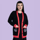 Model wearing Katie Abey x Rebel Romance Pigeon Party Long Cardigan styled over a black dress. Longline black knit with red trim, button front and pocket pigeon details. Alternative, dopamine dressing cardigan available in UK sizes 2XS to 2XL.
