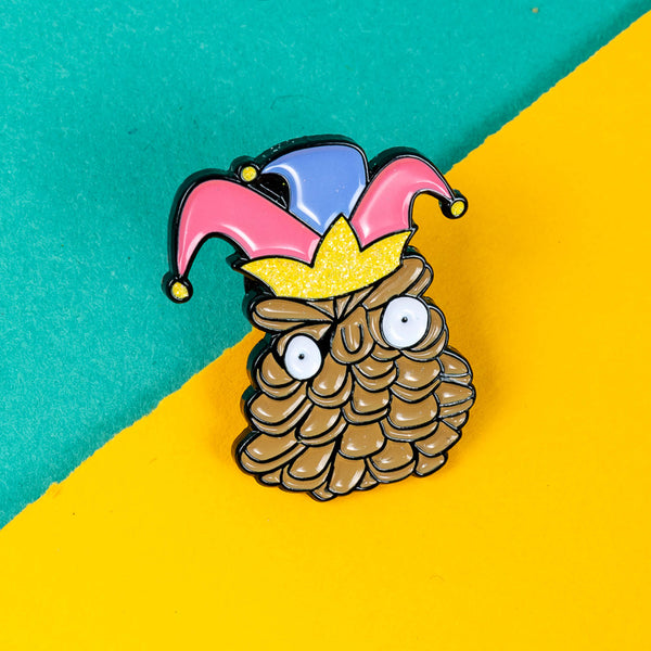 Close-up of the Katie Abey 'Pinecone of Priorities' soft enamel pin. The pin depicts a quirky pinecone character with wide eyes, wearing a vibrant jester hat and crown. Ideal for adding a touch of humour and fun to any outfit or bag.