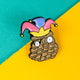 Close-up of the Katie Abey 'Pinecone of Priorities' soft enamel pin. The pin depicts a quirky pinecone character with wide eyes, wearing a vibrant jester hat and crown. Ideal for adding a touch of humour and fun to any outfit or bag.